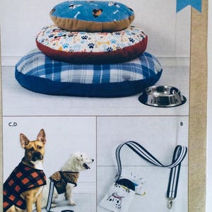 Può includere: Tre cucce per cani impilate, di colore blu, rosso e bianco con motivi. Una ciotola per cani in metallo argentato è su una superficie bianca. Un guinzaglio per cani a strisce bianche e nere con un moschettone in metallo è appeso a una superficie bianca. Un cane marrone e bianco con un giubbotto a quadri rosso e nero è in piedi su una superficie bianca. Un cane bianco con un giubbotto marrone è in piedi su una superficie bianca.