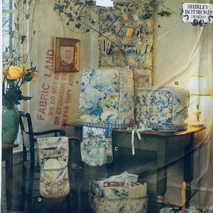 May include: Image of fabric items with a floral design, including chair covers, a basket, and a table cover. The items are arranged on a table and chair, with a lamp and a vase of yellow roses in the background. The text "FABRIC LAND" is visible.