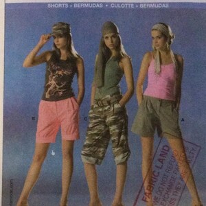 Puede incluir: Tres mujeres modelando diferentes estilos de pantalones cortos. La mujer de la izquierda lleva pantalones cortos rosas con una camiseta de tirantes con estampado gráfico. La mujer del centro lleva pantalones cortos de carga de camuflaje con una camiseta de tirantes verde. La mujer de la derecha lleva pantalones cortos de mezclilla azul claro con una camiseta de tirantes rosa. El texto "FABRIC LAND" es visible en la esquina inferior derecha de la imagen.