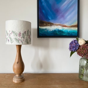 Pu&ograve; includere: Un paralume bianco con un ricamo floreale &egrave; posto su una base di lampada in legno. La lampada &egrave; su un tavolo in legno accanto a un dipinto di un paesaggio marino e un vaso di fiori.
