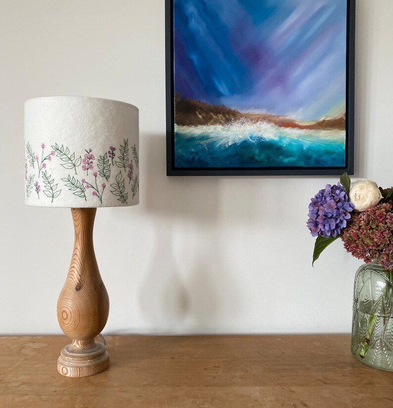 Pu&ograve; includere: Un paralume bianco con un ricamo floreale &egrave; posto su una base di lampada in legno. La lampada &egrave; su un tavolo in legno accanto a un dipinto di un paesaggio marino e un vaso di fiori.