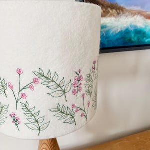 Pu&ograve; includere: Paralume bianco con un ricamo floreale con fiori rosa e foglie verdi.