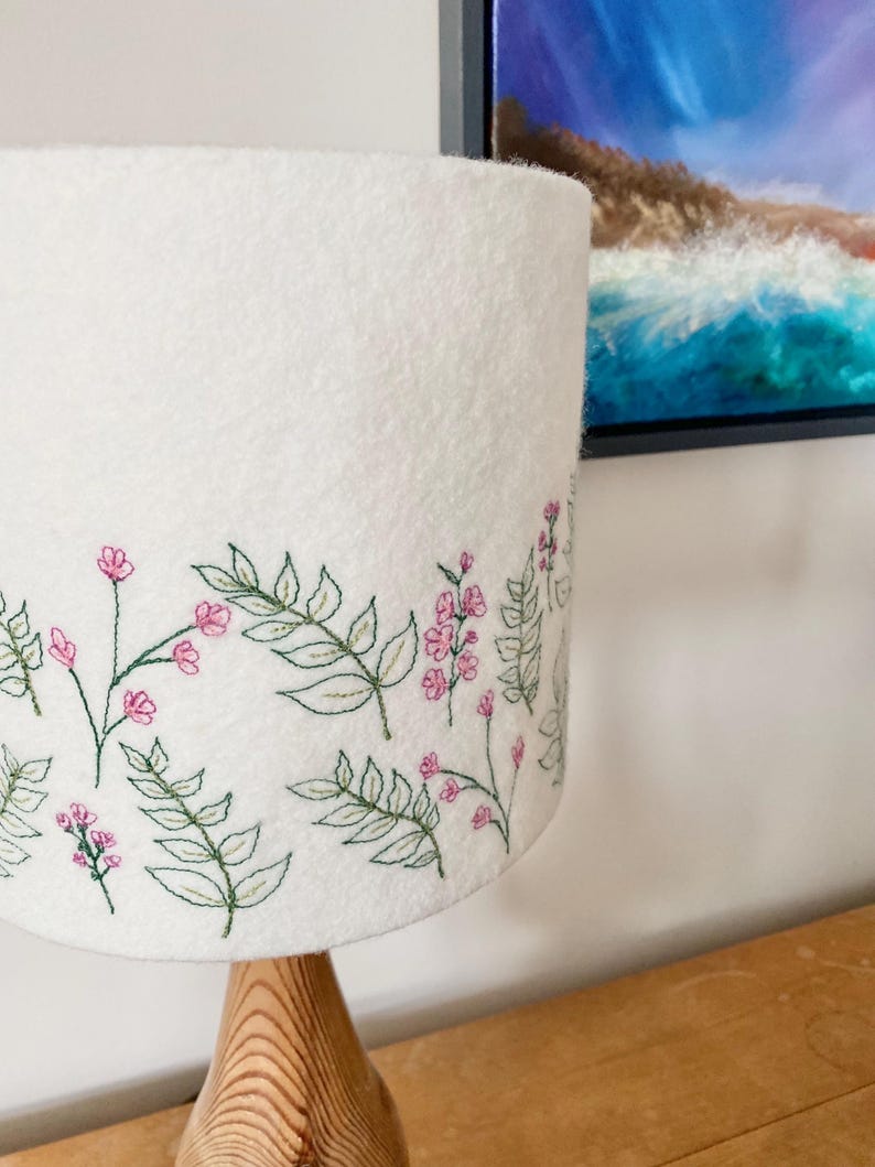 Pu&ograve; includere: Paralume bianco con un ricamo floreale con fiori rosa e foglie verdi.