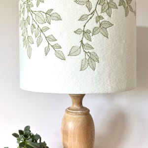 May include: A white lampshade with a green leafy design. The lampshade is on a wooden lamp base.