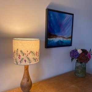 Pu&ograve; includere: Una lampada da tavolo in legno con un paralume bianco con un motivo floreale ricamato. Il paralume presenta fiori rosa e foglie verdi. La lampada &egrave; su un tavolo in legno con un dipinto di un paesaggio marino sul muro dietro di essa.