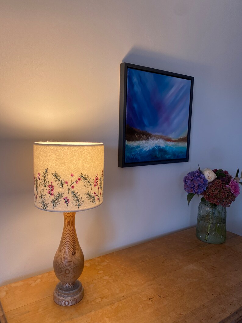 Pu&ograve; includere: Una lampada da tavolo in legno con un paralume bianco con un motivo floreale ricamato. Il paralume presenta fiori rosa e foglie verdi. La lampada &egrave; su un tavolo in legno con un dipinto di un paesaggio marino sul muro dietro di essa.