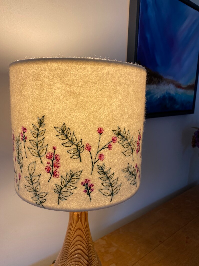 Pu&ograve; includere: Un paralume bianco con un ricamo floreale. Il ricamo presenta fiori rosa e foglie verdi. Il paralume &egrave; su una base di lampada in legno.
