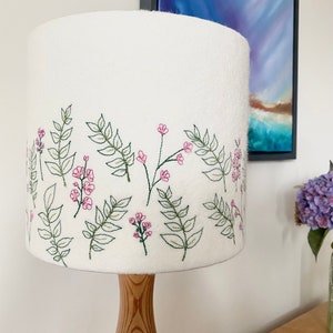 Pu&ograve; includere: Paralume bianco con un ricamo floreale. Il ricamo presenta fiori rosa e foglie verdi.