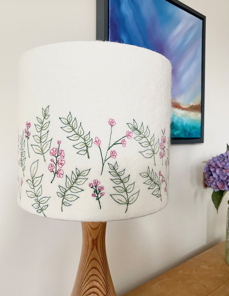Pu&ograve; includere: Paralume bianco con un ricamo floreale. Il ricamo presenta fiori rosa e foglie verdi.