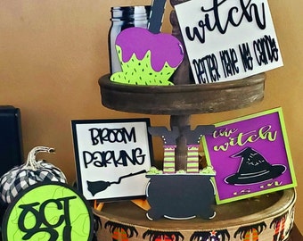 May include: A tiered wooden tray decorated with Halloween-themed decorations. The top tier features a sign that reads "witch better have my candy", a purple and green candy apple, and a mason jar. The middle tier features a sign that reads "broom parking", a black cauldron with green and white striped witch legs, and a sign that reads "the witch". The bottom tier features a green and black circle with the text "oct 31" and a brown burlap ribbon with a repeating pattern of black witch legs.