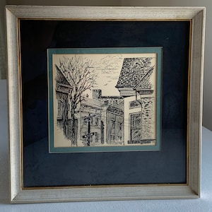 May include: A framed black and white ink drawing of a street scene with buildings and a tree. The drawing is on a cream-colored paper with a blue mat and a black frame.