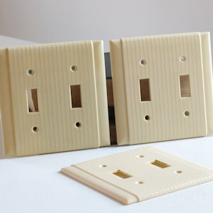 May include: Off-white, vintage-style light switch covers. The covers have a ribbed texture and rectangular openings for the switches. Two double switch plates are standing upright, with a single plate lying flat.