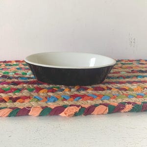 May include: A black and white ceramic bowl sits on a colorful braided mat. The bowl has a dark exterior and a bright white interior. The mat features a variety of colors, including red, orange, green, and blue.