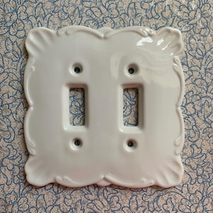 May include: A white ceramic double light switch cover with a decorative, scalloped edge. The cover has two rectangular openings for the switches and four screw holes. The background is a floral pattern.