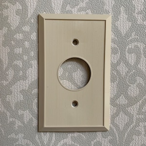 May include: A white plastic electrical outlet cover with a single round opening and two small round holes. The cover has a textured surface.