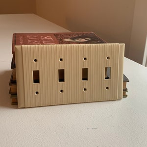 Op de afbeelding: Een crèmekleurige lichtschakelaar met vier schakelaars en verticale ribbels. De afdekking heeft vier rechthoekige openingen voor de schakelaars en vier kleine schroefgaten. Het ligt op een stapel boeken met een bruine kaft.