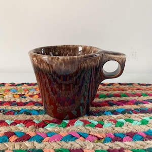 May include: A brown ceramic mug with a unique handle design. The mug has a textured, dripped glaze effect in shades of brown. It sits on a colorful, braided jute placemat with various colors. The mug is designed for hot beverages.
