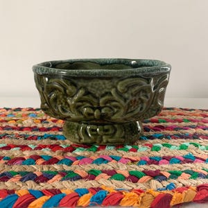 May include: A green ceramic pedestal bowl with a detailed floral design. The bowl has a square shape with rounded corners and sits on a short, round base. The bowl is on a colorful braided mat.