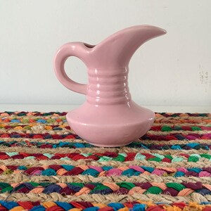 May include: A small, pink ceramic pitcher with a curved spout and a handle. The pitcher has a bulbous base and a ribbed neck. It sits on a colorful braided jute placemat with red, blue, green, and yellow accents.