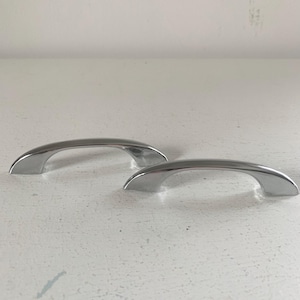 May include: Two polished chrome cabinet pulls with a smooth, arched design. These metal handles feature a reflective, silver-toned finish and a curved shape that tapers at both ends, providing a clean and functional look for furniture or cabinetry.