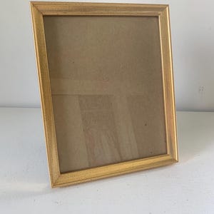 May include: A rectangular picture frame with a metallic gold finish and a textured surface. The frame features a simple, clean-lined design with mitered corners, surrounding a clear glass pane that is set against a neutral, light-colored backing.