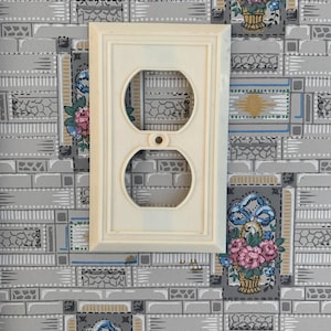 Op de afbeelding: Crèmekleurige stopcontactafdekking met twee ronde openingen en een klein schroefgat. De afdekking heeft een rechthoekig frame met een decoratieve rand. De achtergrond is behang met bloemen- en geometrische patronen.
