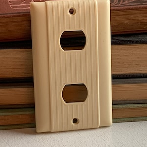 Peut inclure: Plaque de prise électrique vintage de couleur crème avec une texture côtelée verticale. La plaque comporte deux trous de prise et deux trous de vis. La plaque est rectangulaire avec des coins arrondis. Fabriquée en Bakélite ou matériau similaire.
