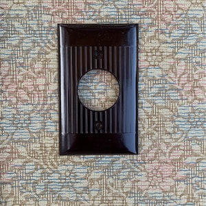 May include: A dark brown electrical socket cover, rectangular with a circular aperture and vertical ribbed detailing. The cover has two screw holes and a glossy finish. The background is patterned fabric with a floral design.