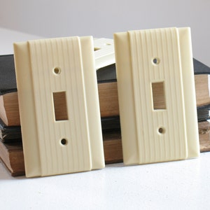 Könnte beinhalten: Cremefarbene Lichtschalterabdeckungen im Vintage-Stil. Die Abdeckungen haben eine rechteckige Form mit einer Öffnung für einen Schalter und zwei Schraubenlöchern. Das Design weist vertikale Rillen auf. Mehrere Abdeckungen sind sichtbar.
