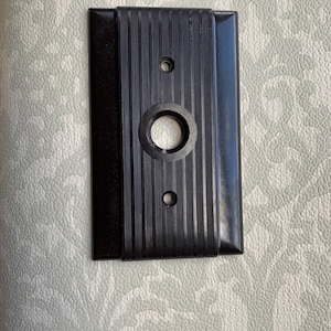 May include: A rectangular, dark brown electrical switch plate with a central circular opening and two smaller holes. The center section has vertical ridged detailing. The plate is likely made of a hard, durable material.