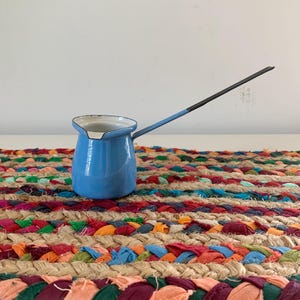 May include: A light blue enamel coffee pot with a long, thin handle. The pot features a white interior, a black rim around the top edge, and a black tip on the end of the handle. The exterior finish is smooth and glossy, showing a solid blue color.