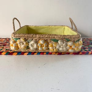Könnte beinhalten: Rechteckiger Weidenkorb mit gelbem Innenfutter und zwei Seilgriffen. Die Vorderseite des Korbs ist mit weißen und gelben Blumenmotiven verziert. Der Korb steht auf einem bunten, gemusterten Textil.