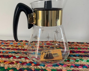 Op de afbeelding: Een vintage glazen koffiekan met een zwarte handgreep en deksel, en een goudkleurige band. De kan heeft een heldere body met sterrenpatronen. Een kleine kaart met tekst zit erin. De kan staat op een kleurrijke geweven mat.