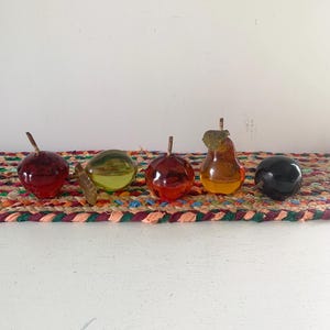 May include: Five glass fruit ornaments in various colors: red apple, green apple, red apple, yellow pear, and black plum. Each piece has a small brown stem. The fruits are arranged on a colorful woven mat.
