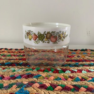 May include: A clear glass Pyrex container with a white lid featuring a colorful fruit and vegetable design. The container sits on a multi-colored braided placemat, adding a touch of vintage charm to any kitchen.
