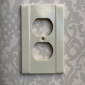 Könnte beinhalten: Cremefarbene Steckdosenabdeckung mit zwei kreisförmigen Öffnungen und einem kleinen zentralen Loch. Die rechteckige Abdeckung hat eine leicht glänzende Oberfläche. Das Design ist schlicht und funktional.