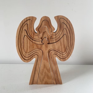 May include: A wooden angel sculpture with a layered design. The sculpture is made of light brown wood and features a detailed outline of an angel with outstretched wings. The angel stands upright on a flat base.