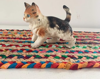 May include: A ceramic figurine of a kitten with orange, black, and white markings. The cat has blue eyes and is posed in a walking position. It is placed on a colorful braided rug with red, blue, and orange hues.