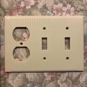 May include: Off-white electrical wall plate with one outlet and two switch openings. The plate has a vertical ribbed texture. The background is a floral patterned fabric.
