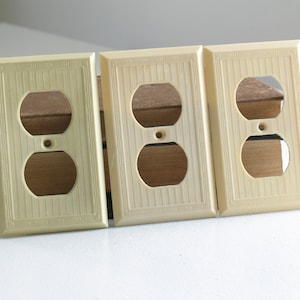 May include: Three off-white electrical outlet covers. Each cover has a rectangular shape with a textured surface and a decorative border. The covers have two circular openings for the outlets and a small hole for a screw.