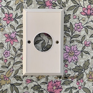 May include: A cream-coloured electrical outlet cover with a circular opening and two screw holes. The cover has a textured, vertically-lined surface and a rectangular frame. The background features floral wallpaper with pink and yellow flowers.