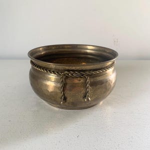 May include: A round brass bowl featuring a polished metallic finish and a decorative rope-style trim encircling the upper exterior. The bowl has a wide, open top and a smooth, curved body, showcasing a warm, golden-brown patina throughout the surface.