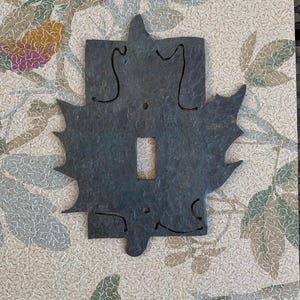 Peut inclure: Un cache-interrupteur décoratif en métal gris foncé avec un motif de feuille. Le couvercle comporte une ouverture centrale rectangulaire pour l'interrupteur et est entouré d'une forme de feuille stylisée avec des bords pointus. Le fond présente un motif floral.