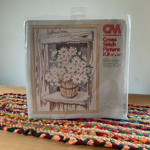 Op de afbeelding: Een kruissteekpakket van Columbia-Minerva met een bloemenontwerp. De kit bevat een gestempeld ontwerp op 100% katoenen stof, met een afmeting van 30.5 cm x 40.6 cm. De verpakking is van helder plastic.