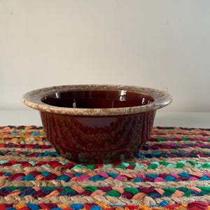 May include: A brown ceramic bowl with a glossy finish and a lighter-colored rim. The bowl sits on a colorful braided placemat with various hues of red, blue, green, and orange.