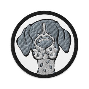 May include: A circular embroidered patch featuring a stylized dog head with grey fur, a white muzzle with grey spots, and floppy ears. The design is set against a white background and is framed by a thick, solid black stitched border edge.