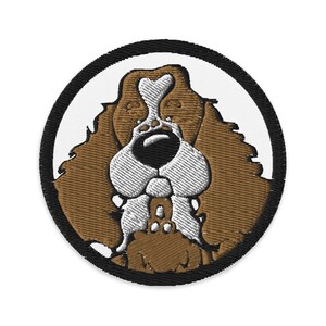 May include: Embroidered patch of a brown and white cartoon dog with a black nose and big eyes. The dog is inside a black circle with a white background.
