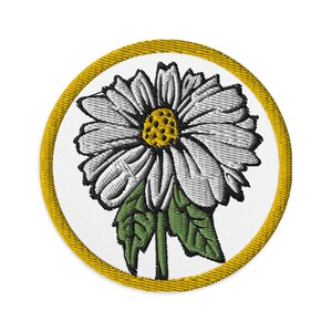 Op de afbeelding: Een witte madeliefje met een gele kern en groene bladeren is geborduurd op een witte cirkel met een gouden rand.