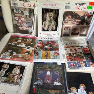 May include: A selection of craft pattern packets with festive themes. The packets display designs for Christmas and holiday decorations, including snowmen, angels, and Santa Claus figures. The packets are in various sizes and colors, with text such as "Simplicity Craft" and "McCall's Crafts".