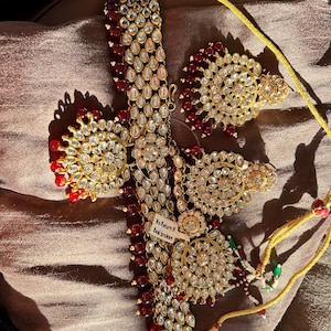 Puede incluir: Un conjunto de collar y pendientes dorados con piedras preciosas claras y rojas. El collar presenta un colgante central con un diseño floral, y los pendientes tienen un patrón circular similar. El conjunto se exhibe sobre una superficie marrón texturizada.